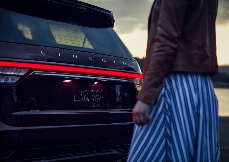 A person is shown near the rear of a 2023 Lincoln Aviator® SUV as the Lincoln Embrace illuminates the rear lights | Dave Sinclair Lincoln in Ballwin MO