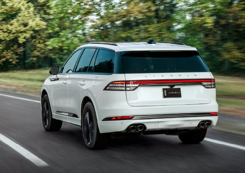 A white 2026 Lincoln Aviator® SUV is being driven away down a road lined with colorful trees. | Dave Sinclair Lincoln in Ballwin MO