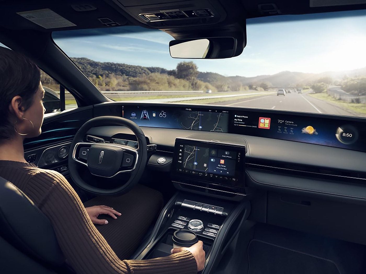 Woman driving a Lincoln Nautilus with the wide digital display and a sunny hillside road ahead