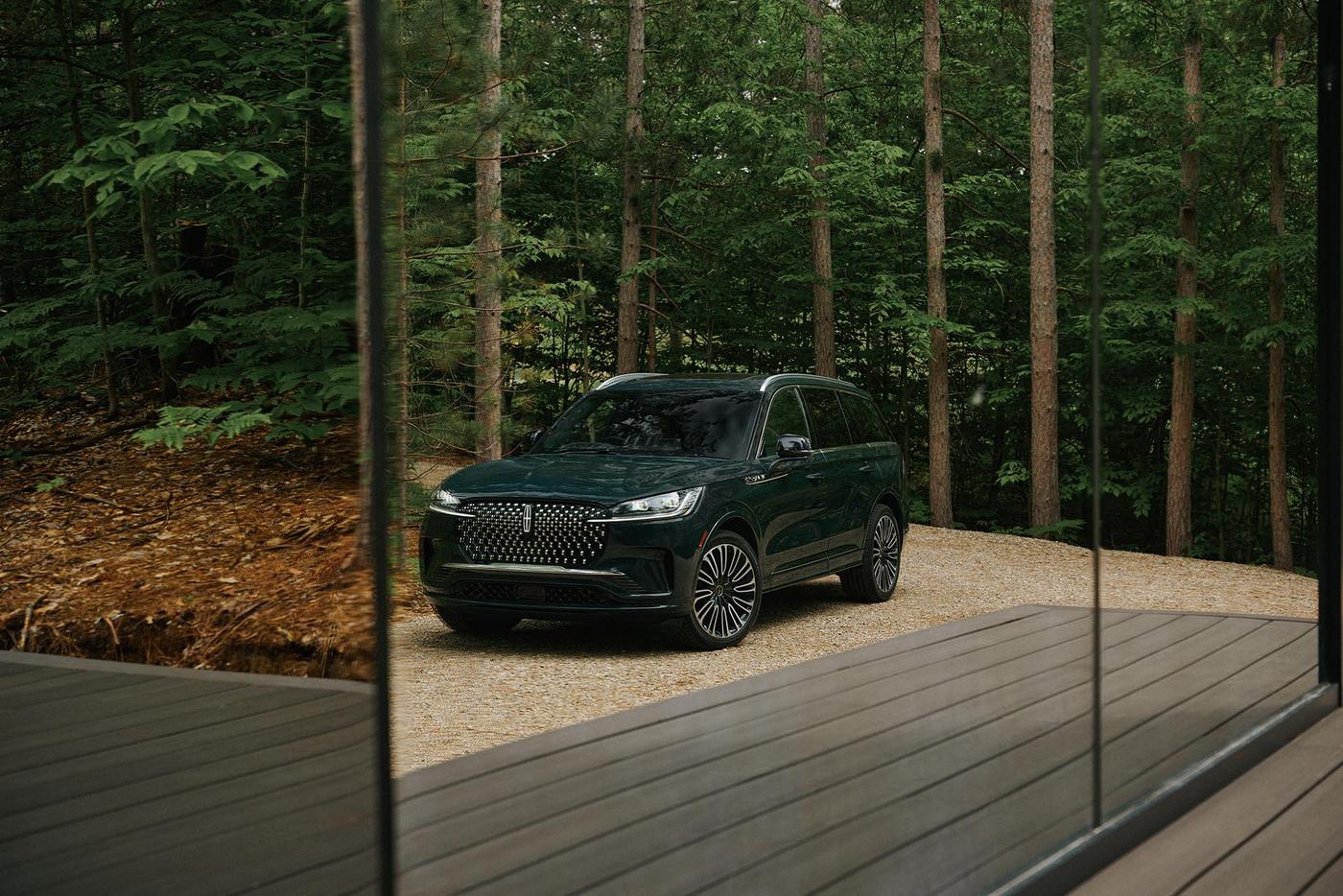 Dark green Lincoln Aviator parked on gravel beside a glass wall in a forest